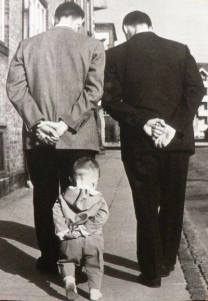 Fotografía en blanco y negro: un niño pequeño, de espaldas, camina detrás de dos hombres imitándolos con las manos a la espalda
